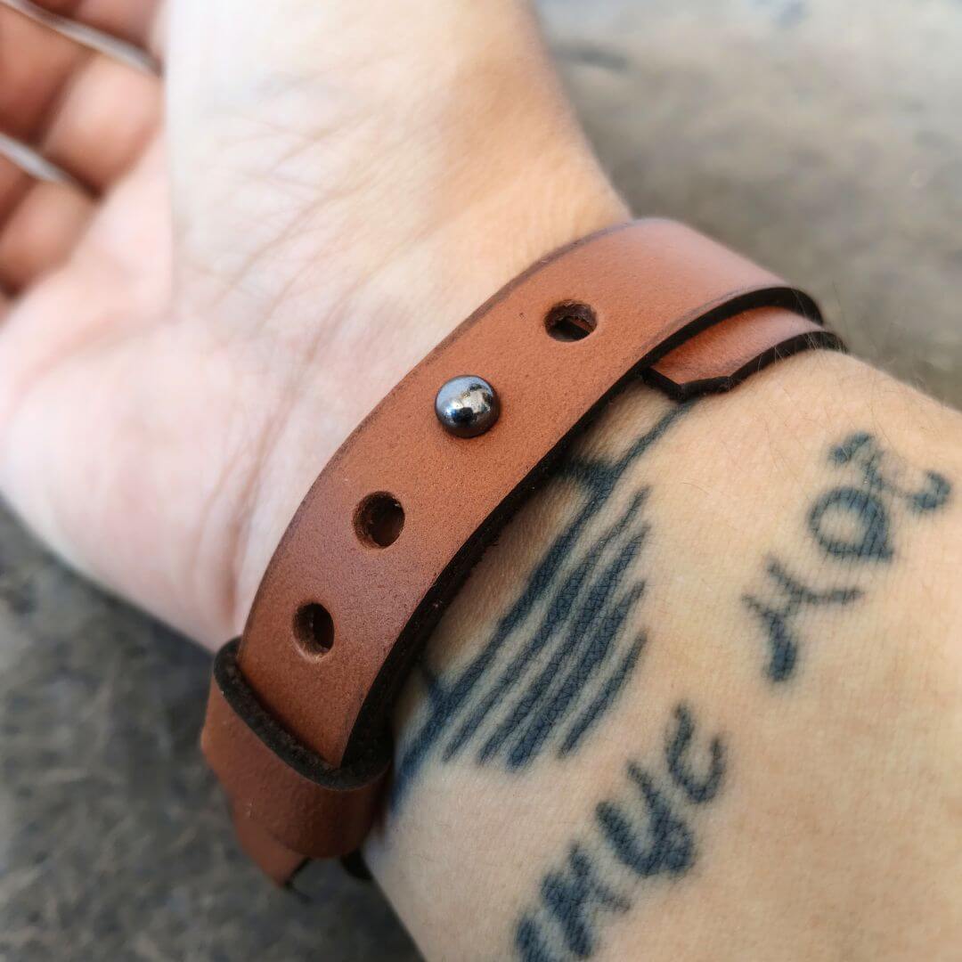 A close-up of a tan leather wristband worn on a person's wrist, featuring a silver metal rivet and adjustable holes.