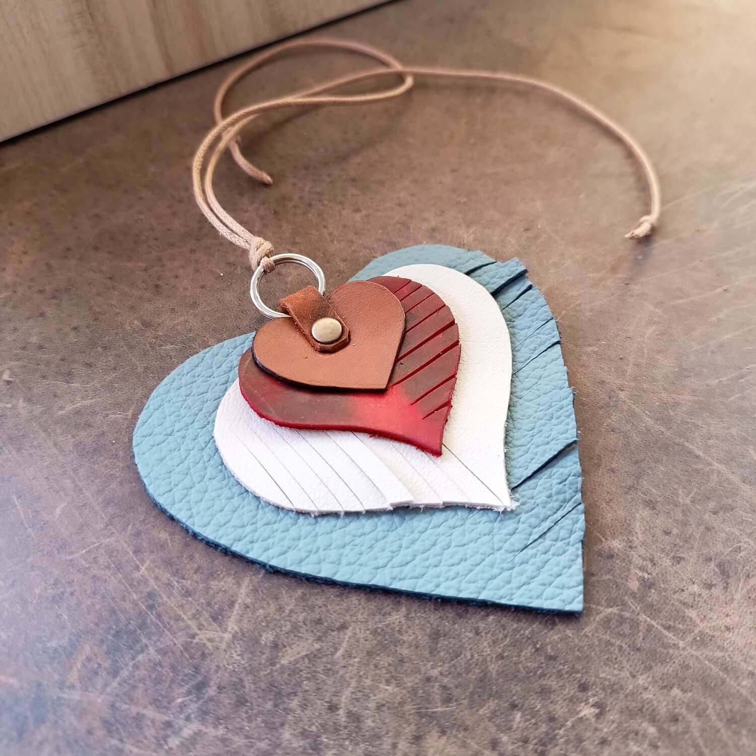 Angled view of The "Heartstrings" Charm, illustrating the depth of its four layered leather hearts, the distinct fringe patterns, and the secure leather loop and rivet attachment.