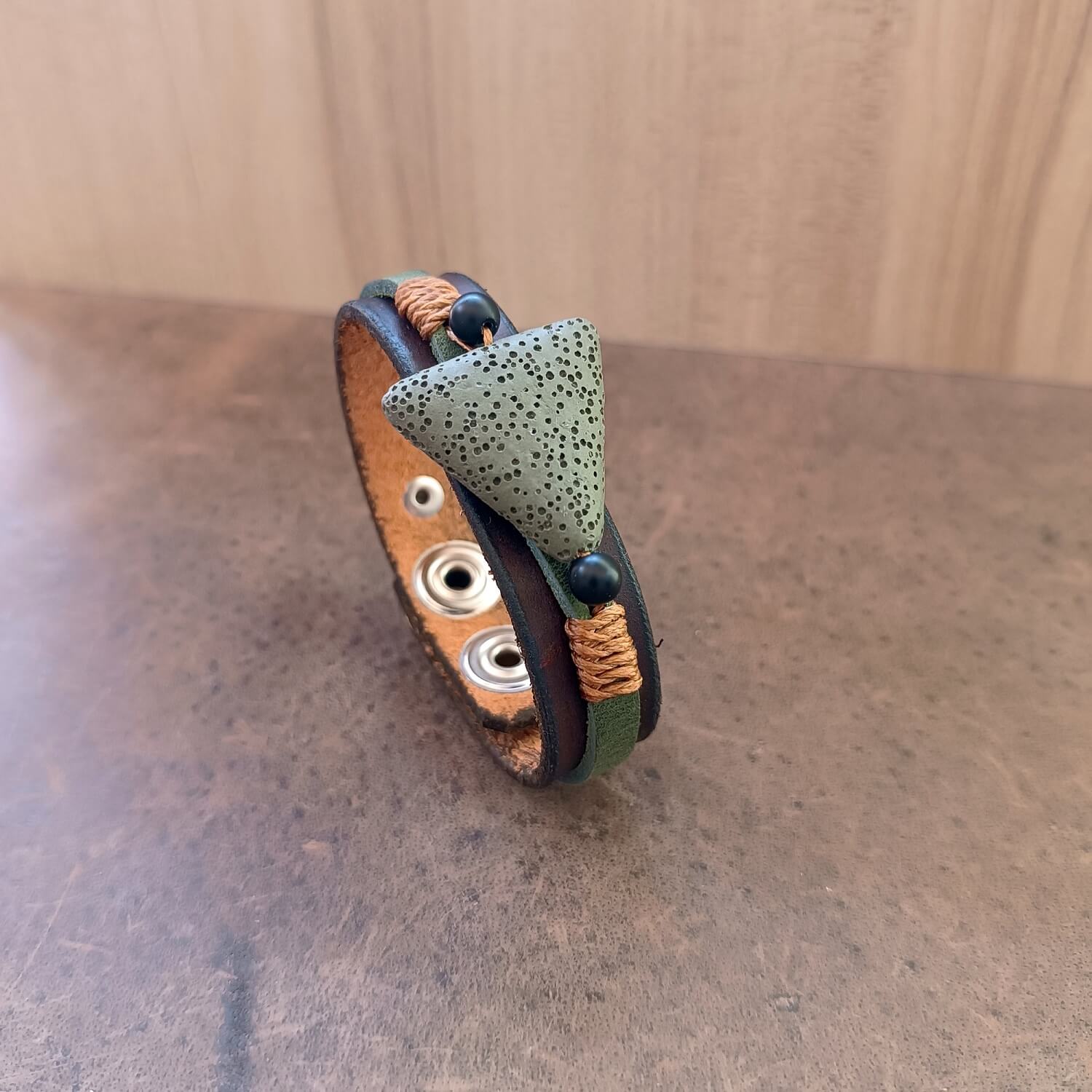 The Earth's Pulse Aromatherapy Bracelet with Green Lava. Shows the wide dark brown Crazy Horse leather band, a khaki green porous lava bead flanked by matte black onyx beads, and bronze waxed cord.