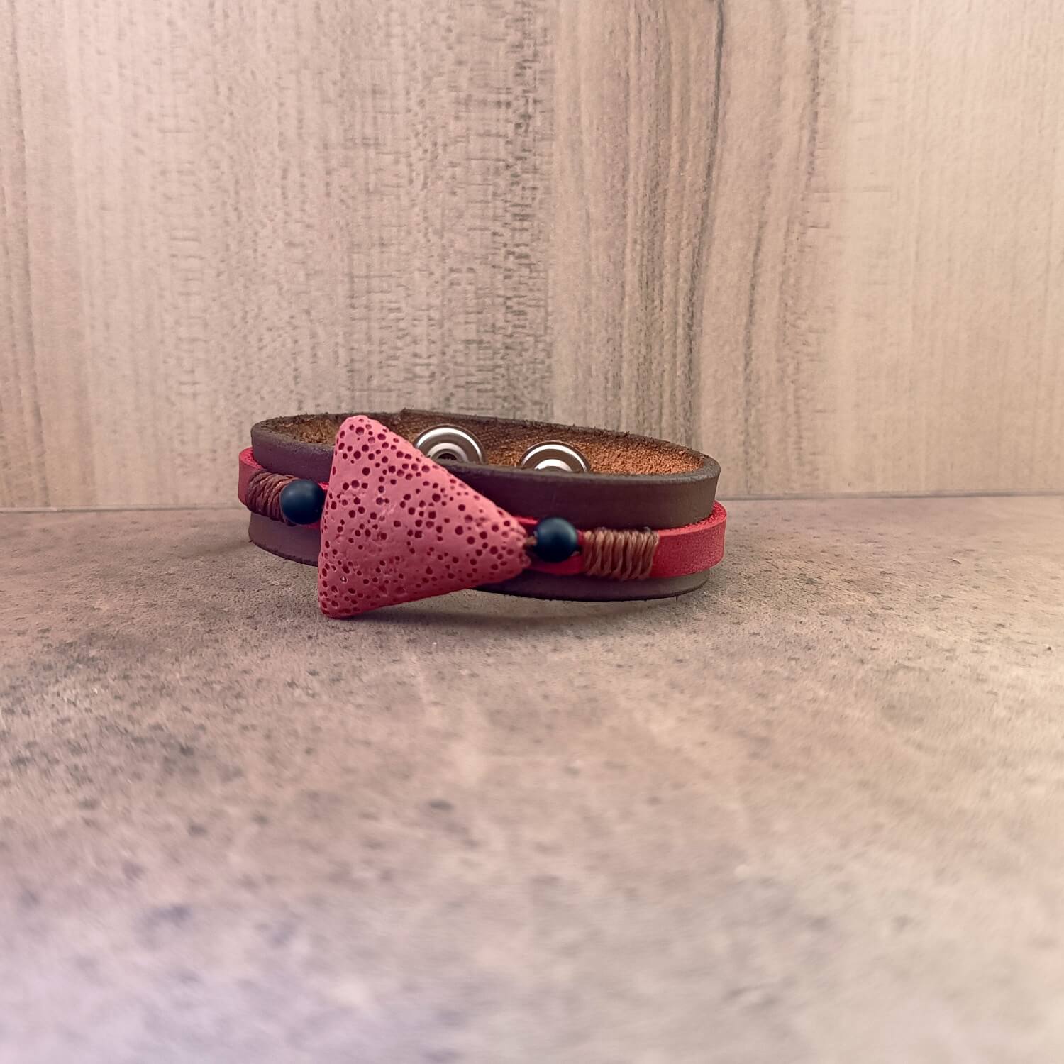 The Earth's Pulse Aromatherapy Bracelet with Red Lava, laid flat. Shows the wide dark brown Crazy Horse leather band, a brick red porous lava bead flanked by matte black onyx beads, and bronze waxed cord.