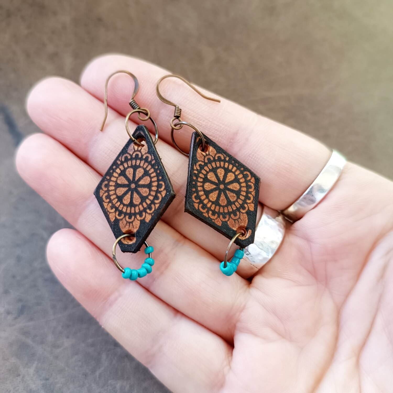 A pair of boho style dangle earrings with a geometric mandala design, featuring turquoise czech beads and antique bronze hooks, held in a person's hand.