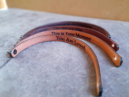 Back of Personalized Thin Leather Bracelets in Mustard Brown and Dark Brown variations, showing custom engraved text (Displayed text: " You are loved", "This is your moment", "one step at a time") on the underside.