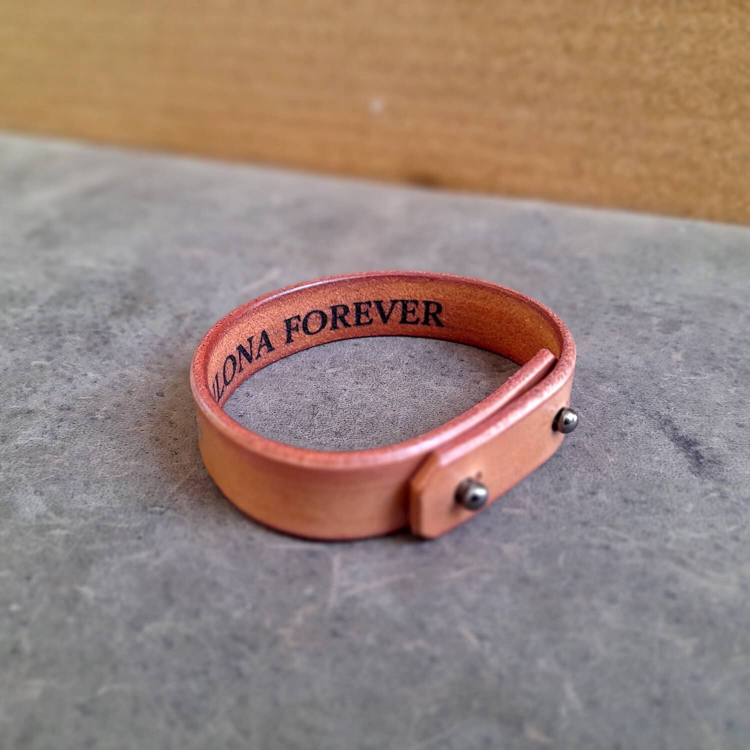 Brown leather bracelet with 'LONA FOREVER' engraving on a gray surface