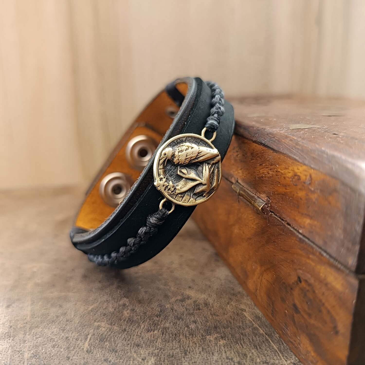 Black braided leather bracelet with a brass raven emblem on a wooden surface