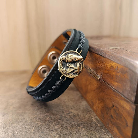 Black braided leather bracelet with a brass raven emblem on a wooden surface