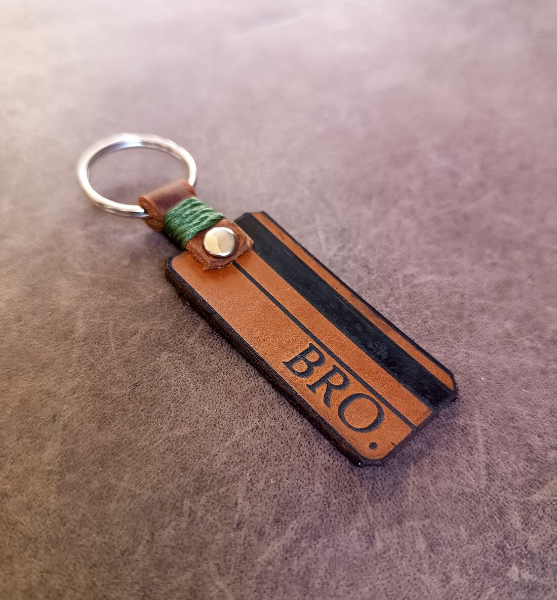 A personalized brown leather keychain with the text 'BRO' engraved on it, featuring a silver keyring and green thread detail.