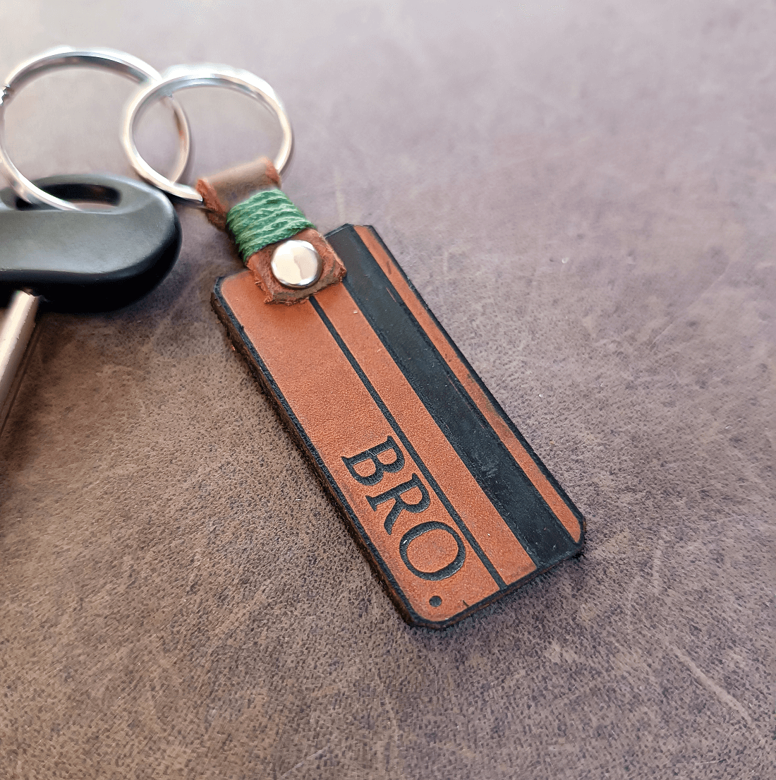 A personalized brown leather keychain with the text 'BRO' engraved on it, featuring a silver keyring and green thread detail.