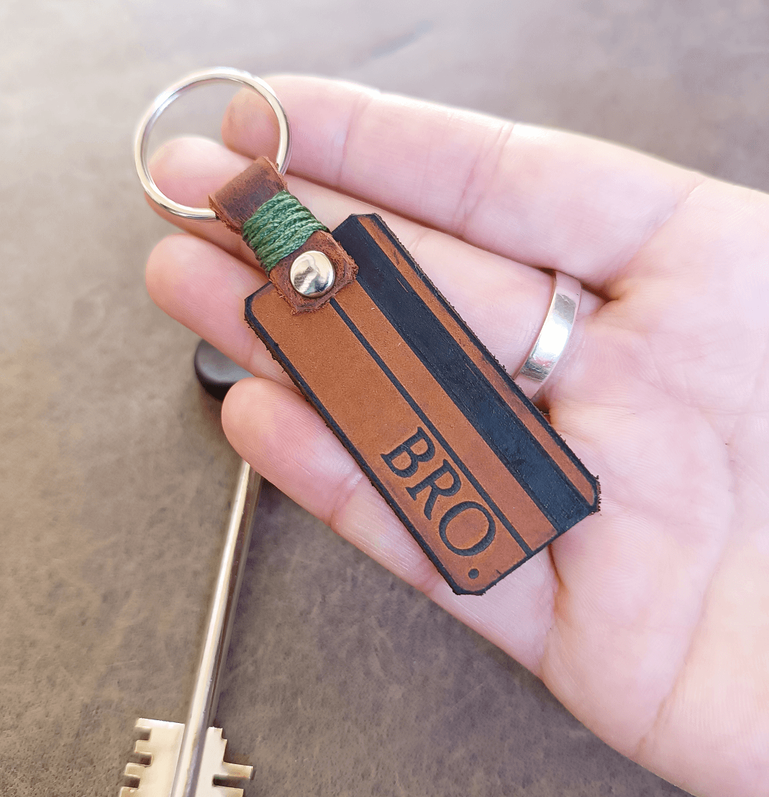 Hand holding a leather keychain with 'BRO.' engraving on a wooden surface