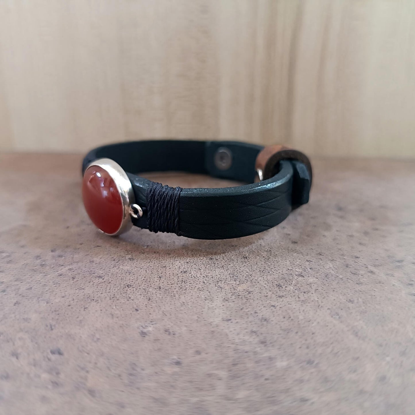 Black leather bracelet with a red stone and silver clasp on a textured surface.
