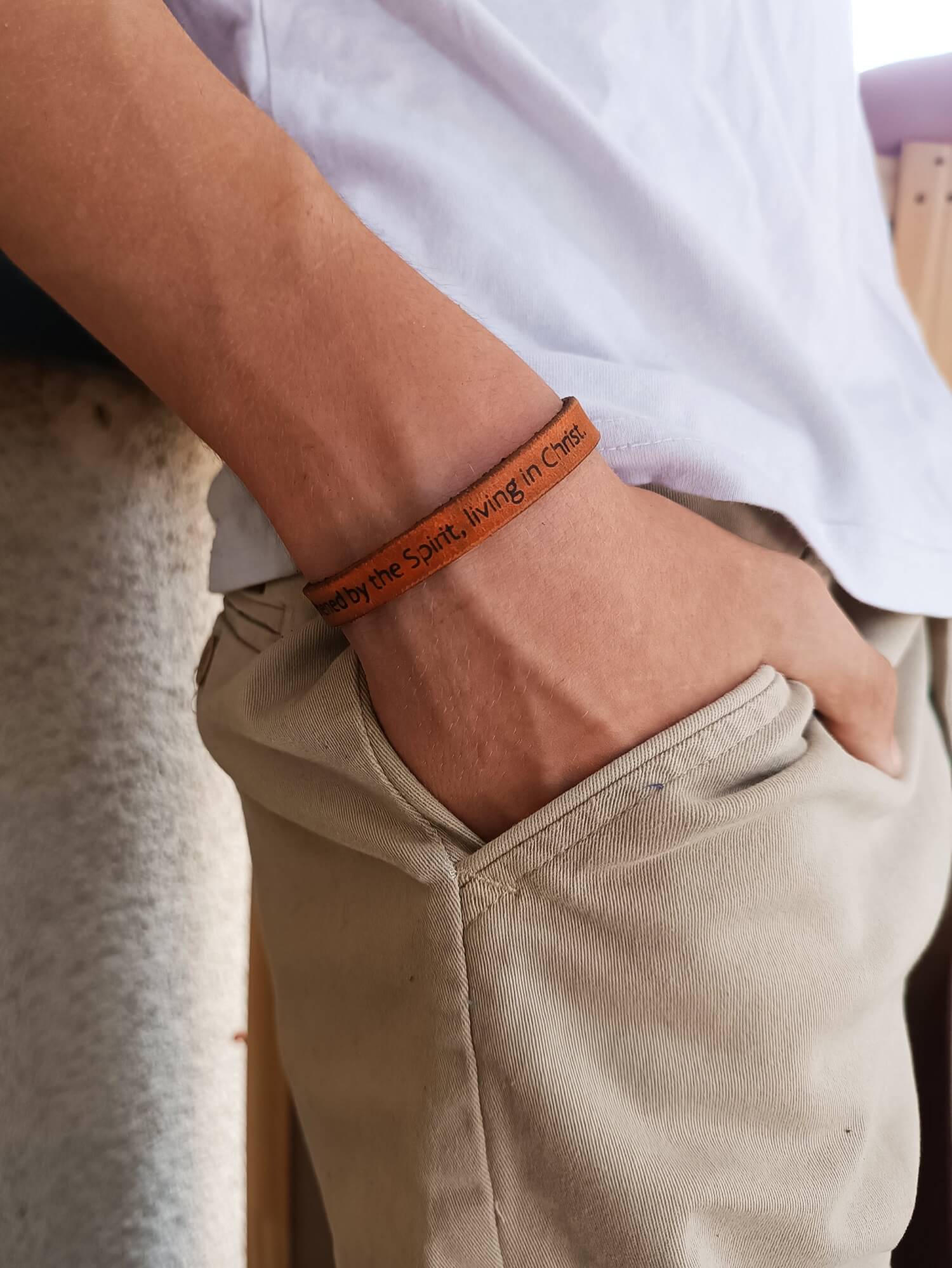 Person wearing a brown leather bracelet with text on a white shirt and beige pants.