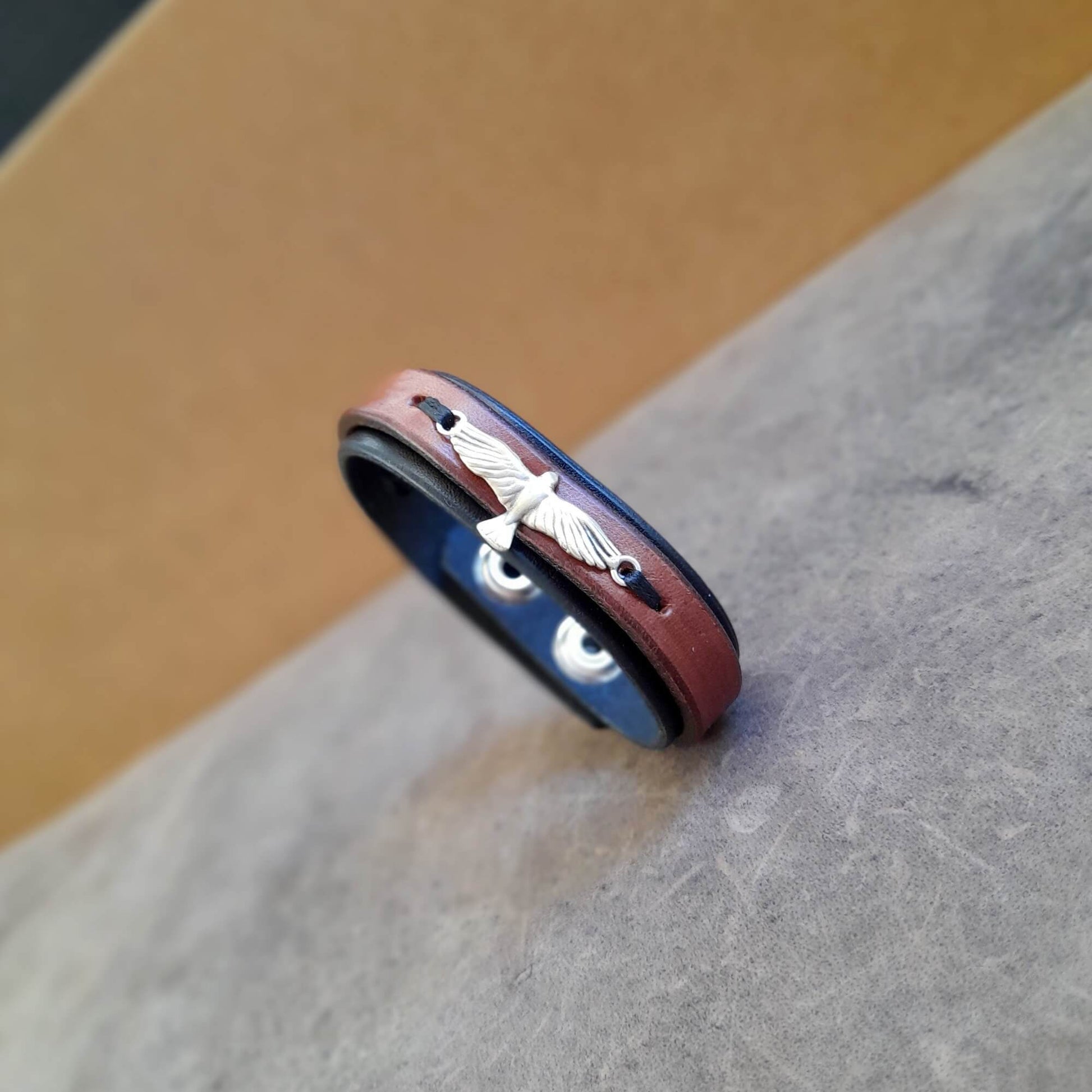 A layered black and brown leather bracelet with an eagle emblem on the top, displayed on a textured background.
