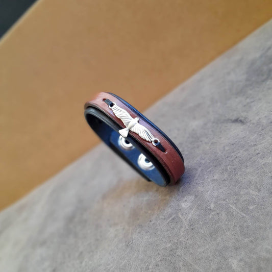 A layered black and brown leather bracelet with an eagle emblem on the top, displayed on a textured background.