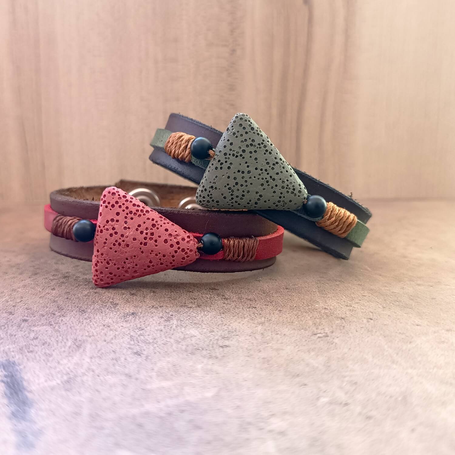 The Earth's Pulse Aromatherapy Bracelets duo. Shows the wide dark brown Crazy Horse leather band, a khaki green and brick red porous lava bead flanked by matte black onyx beads, and bronze waxed cord.