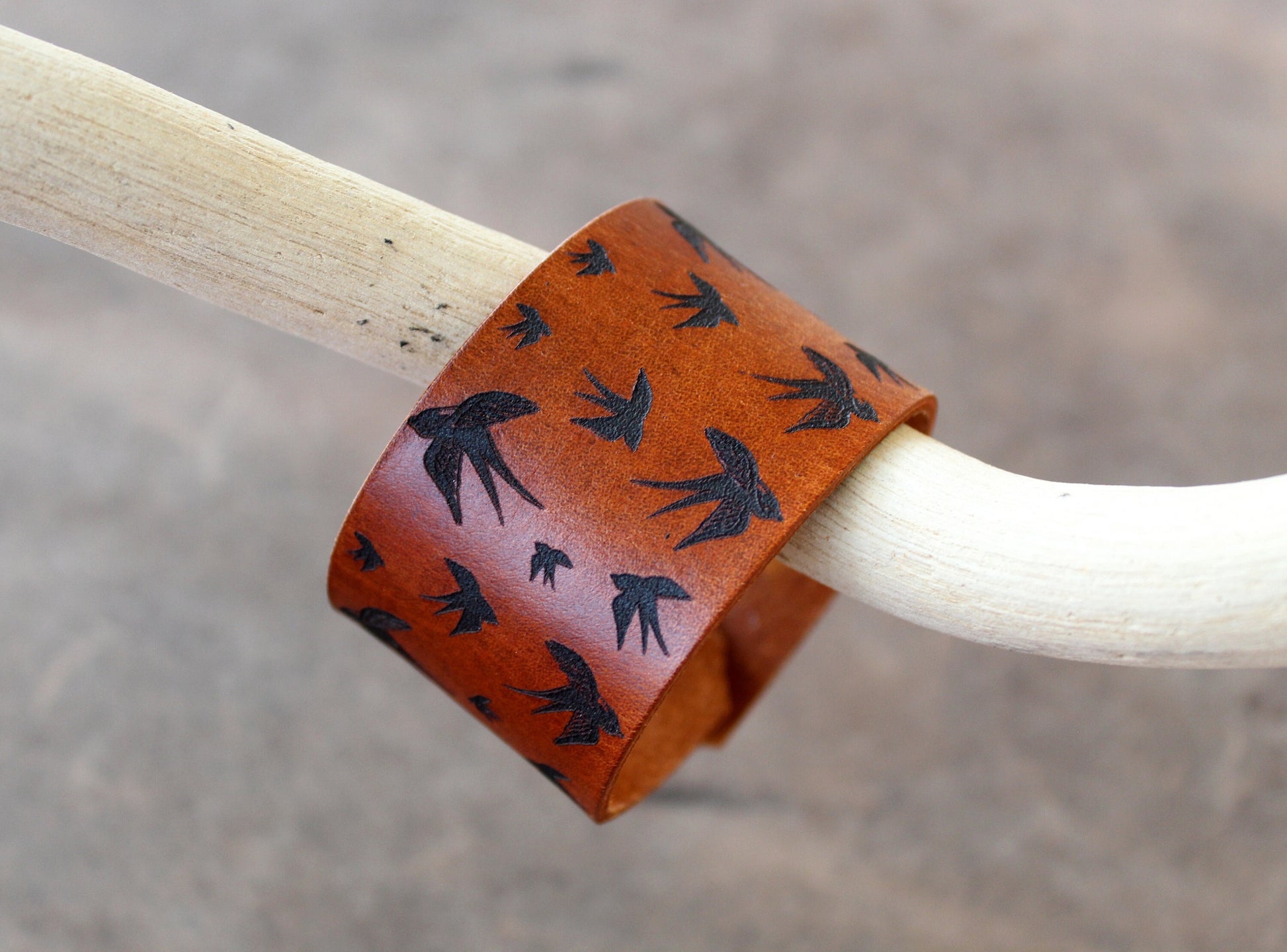 Flying Birds Wide Leather Cuff Bracelet on warm brown genuine leather, featuring a deep hand-engraved design of birds in flight, hanging sideways from driftwood.