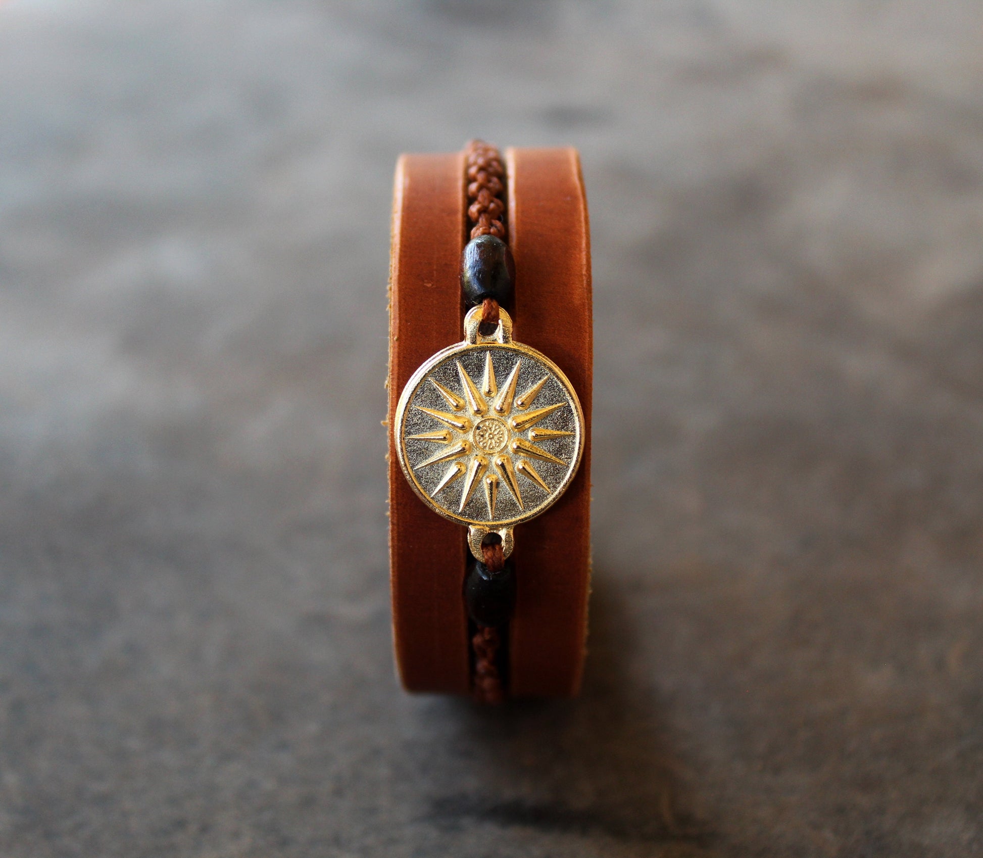 Gold Vergina Sun Leather Bracelet with gold-plated Vergina Sun & Alexander the Great charm on flat surface. Features brown leather, wooden beads, and macrame.