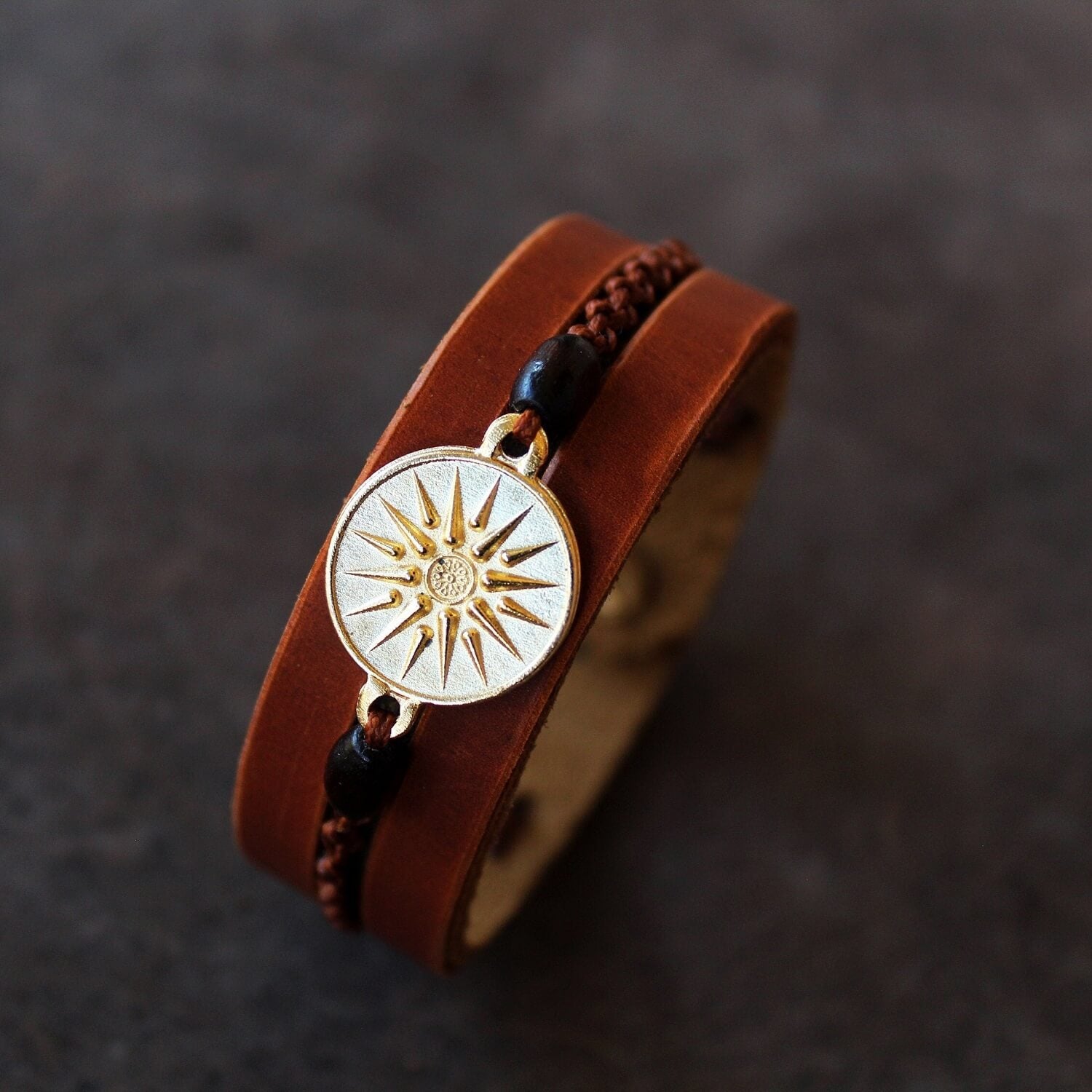 Gold Vergina Sun Leather Bracelet with gold-plated Vergina Sun & Alexander the Great charm on flat surface. Features brown leather, wooden beads, and macrame.