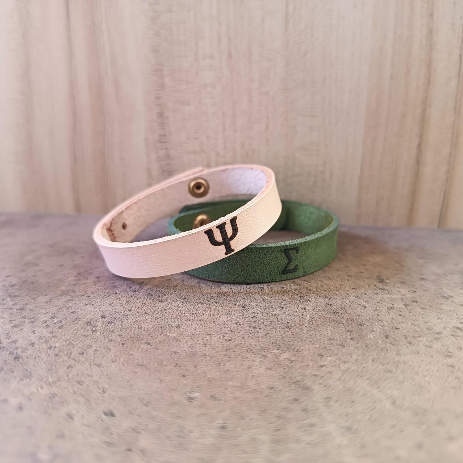 Two leather bracelets with Greek letters engraved on them, one in natural color and the other in green, displayed on a wooden surface.