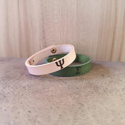 Two leather bracelets with Greek letters engraved on them, one in natural color and the other in green, displayed on a wooden surface.