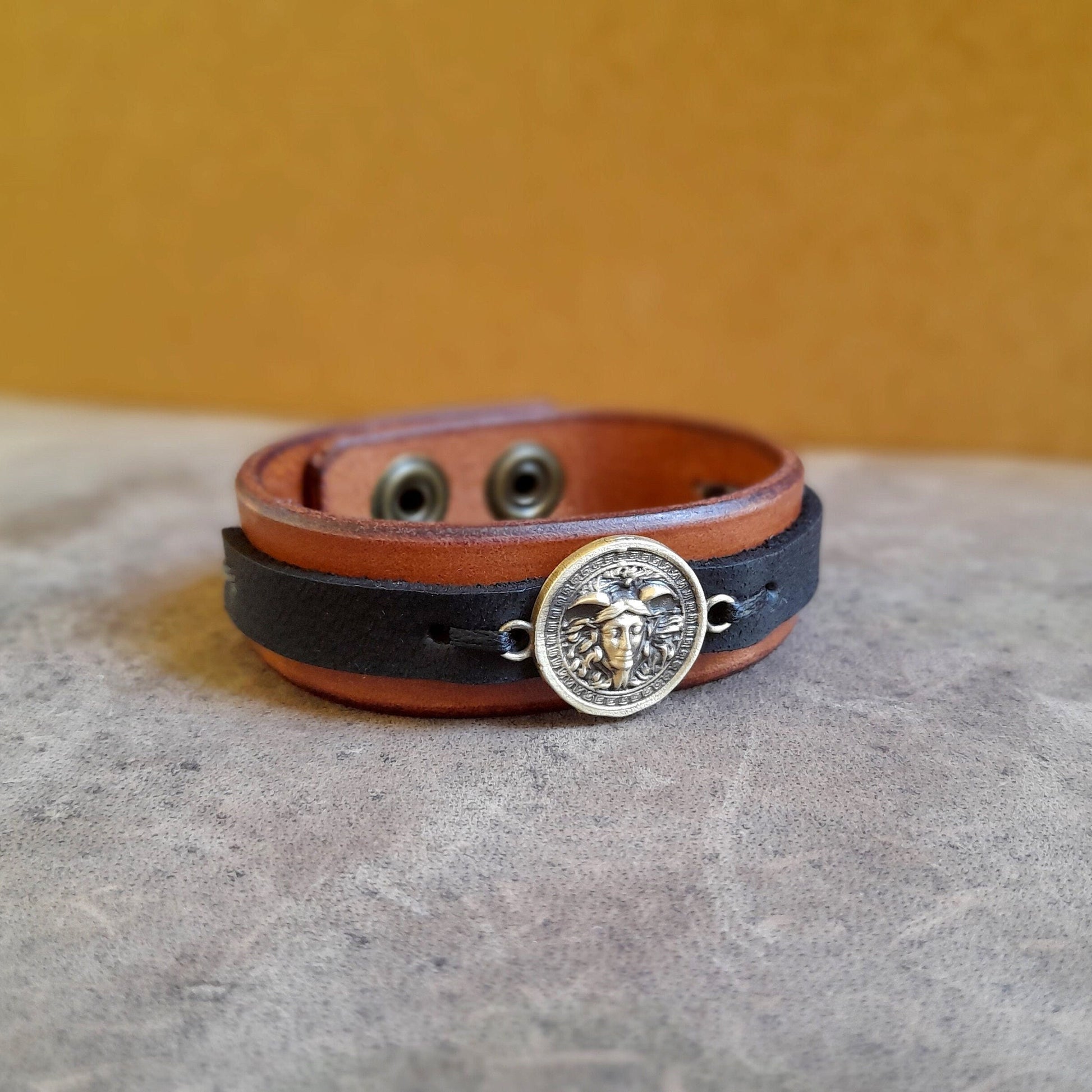 Greek Medusa Leather Bracelet on flat surface, showing handcrafted oxidized brass Medusa charm, brown Crazy Horse leather, and black stripe.