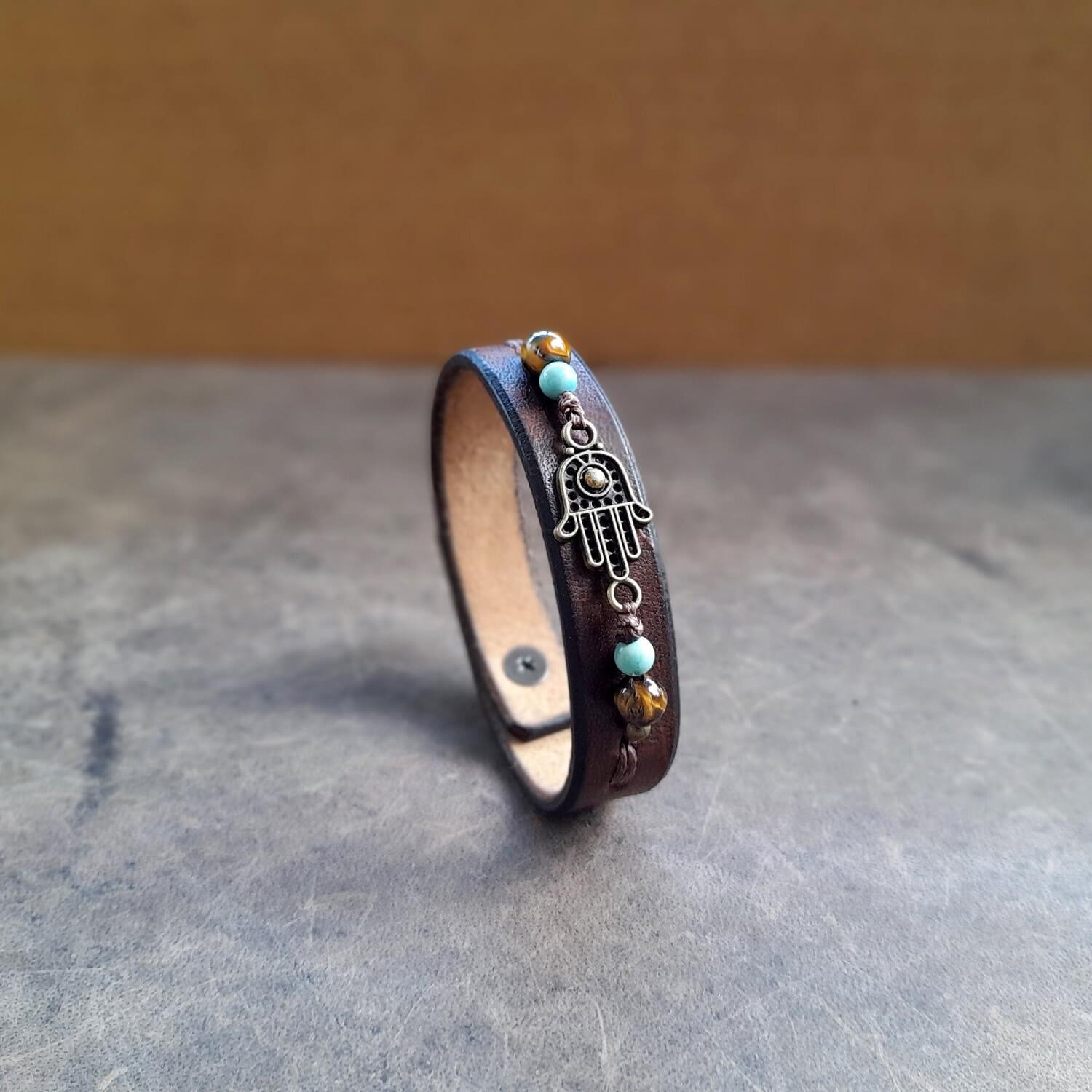 The Hamsa Hand Beaded Leather Bracelet: A chestnut brown full-grain leather cuff featuring a central Hamsa Hand flanked by green Turquoise and Tiger's Eye gemstone beads.