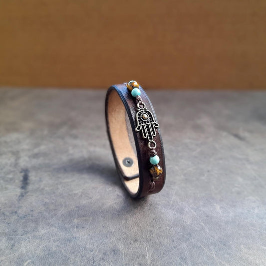 The Hamsa Hand Beaded Leather Bracelet: A chestnut brown full-grain leather cuff featuring a central Hamsa Hand flanked by green Turquoise and Tiger's Eye gemstone beads.