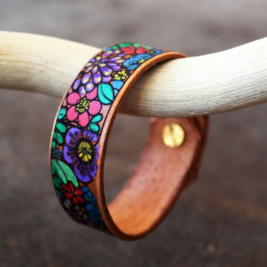 Hand-Painted Flowers Bracelet on driftwood, showing vibrant floral pattern inspired by stained glass art. Features purple, lilac, turquoise, yellow, pink, red, green tones on leather.
