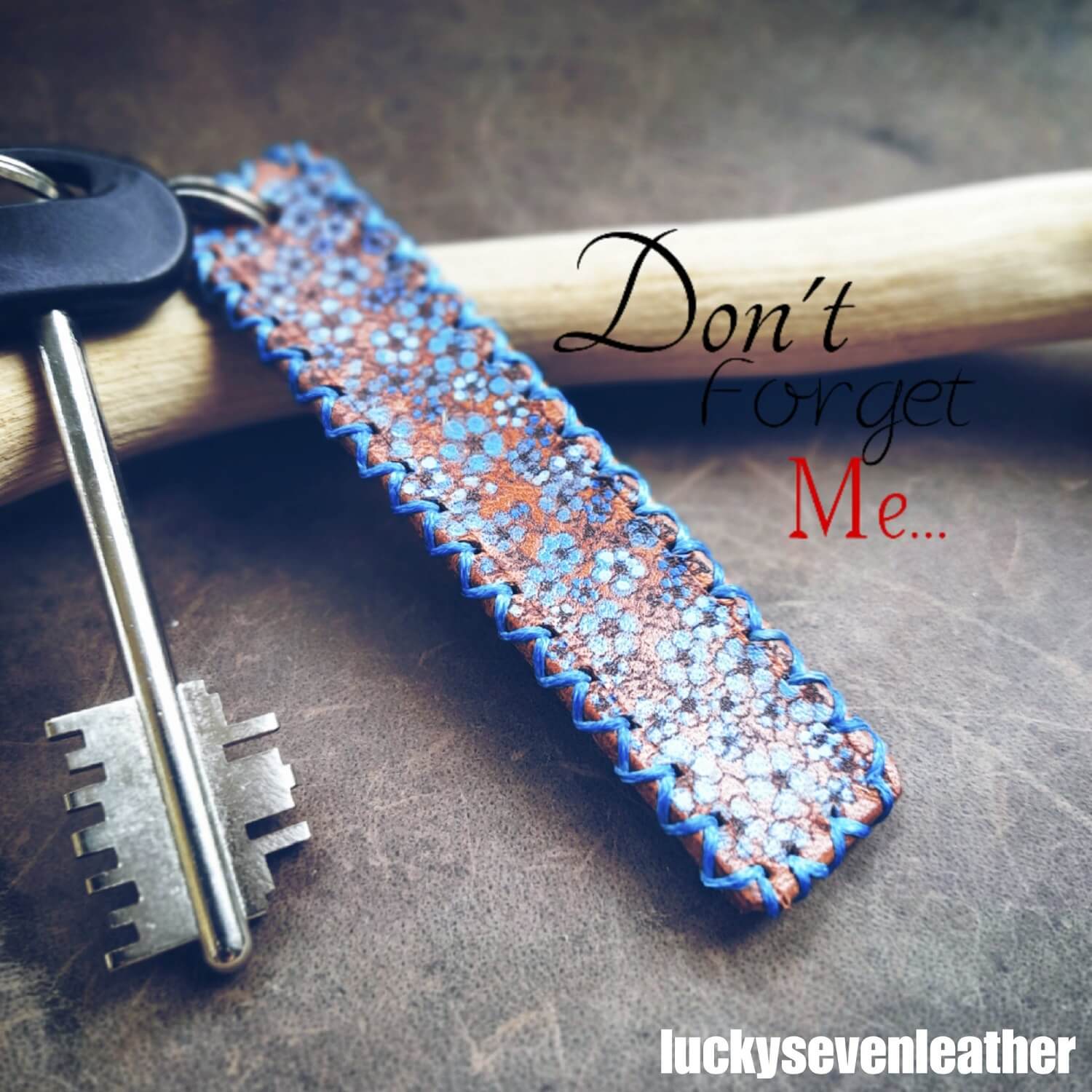 The Hand Painted Forget Me Not Personalized Leather Keychain on a beautiful poster with caption "Don't forget me": A brown Crazy Horse leather keychain featuring delicate hand-painted forget-me-not flowers, blue hand-stitching, and two split rings.
