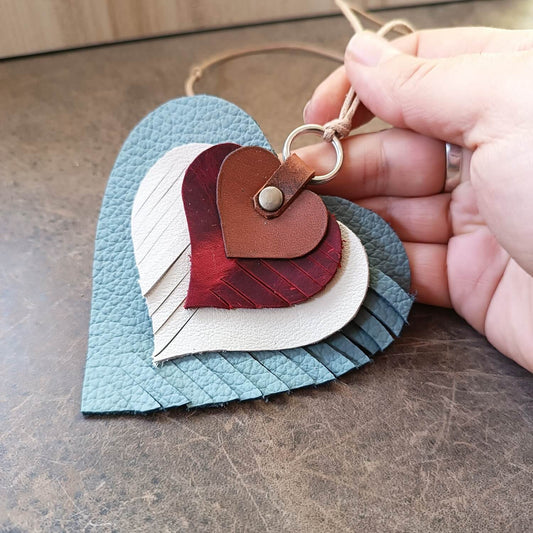 Hand holding The "Heartstrings" Charm: a unique, four-layered heart ornament crafted from upcycled leather remnants in brown, burgundy red, white, and light blue, with rotating fringe patterns and a cotton cord.