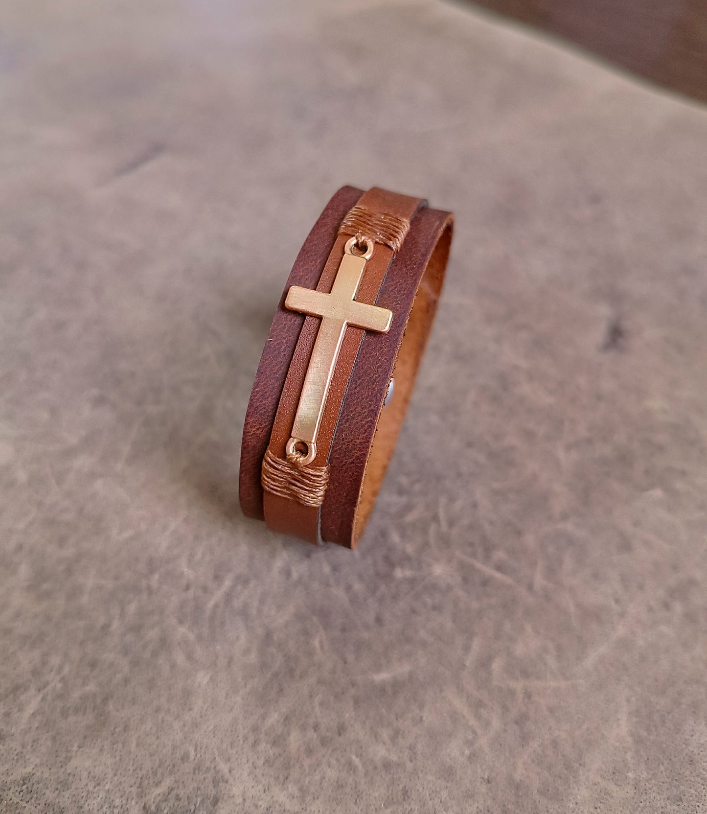 Layered brown leather cuff with rose gold cross charm and customizable underside for engraving Bible verses or personal messages.