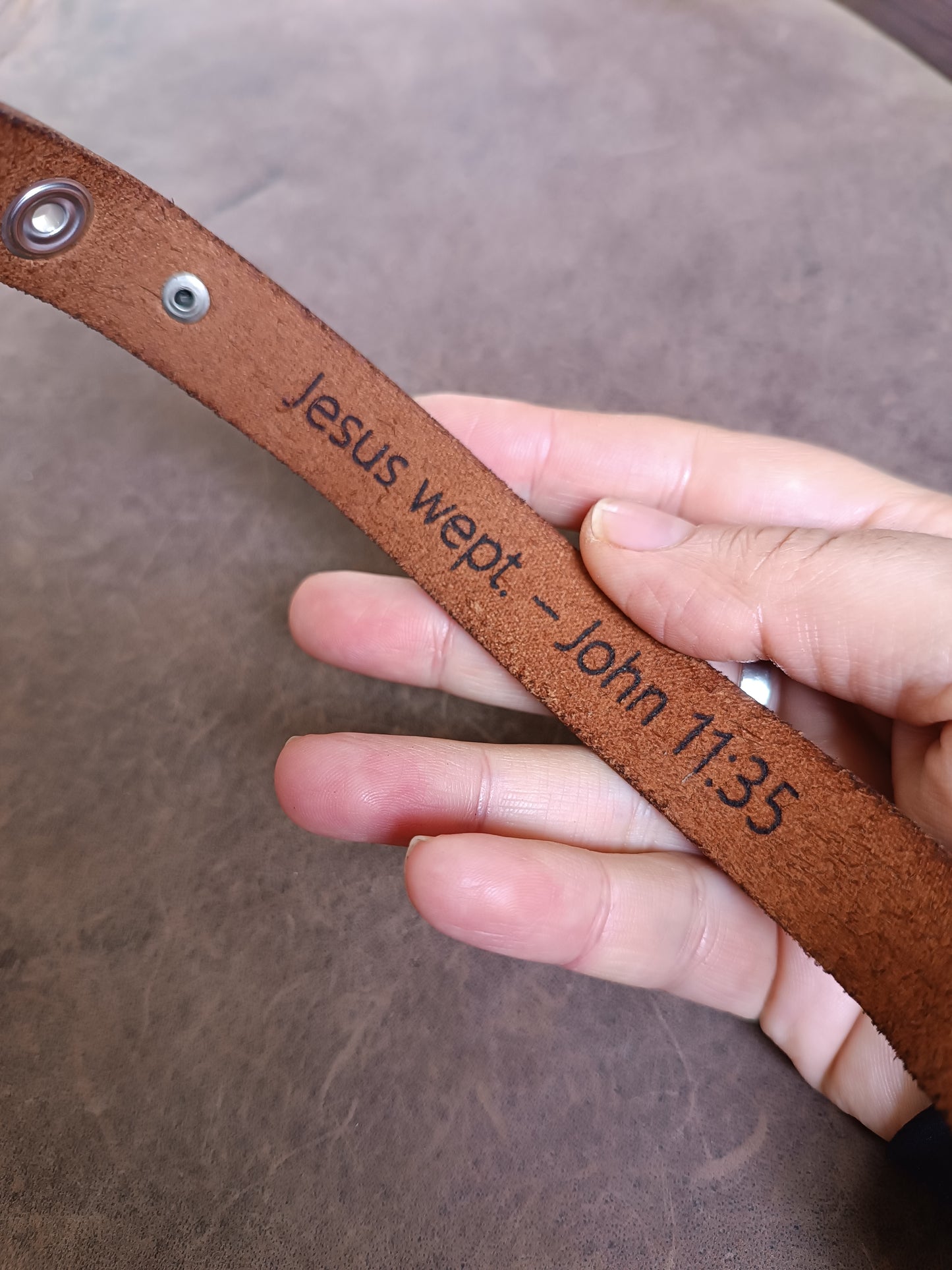 Layered brown leather cuff with rose gold cross charm and customizable underside for engraving Bible verses or personal messages.