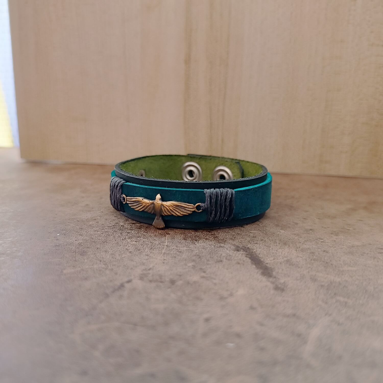 Front view of the Inzaghi Bracelet showing the double-layered black/green leather base with a muted dark petrol blue top strip. The oxidized brass dove pendant, wings open and ready to soar, sits centered amid five industrial gray waxed cord wraps symbolizing silent loyalty and steadfast presence.