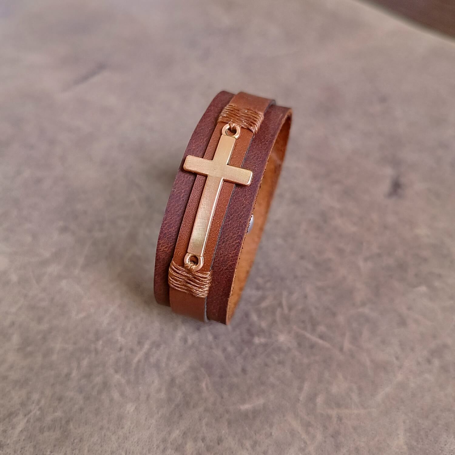 Layered brown leather cuff with rose gold cross charm and customizable underside for engraving Bible verses or personal messages.