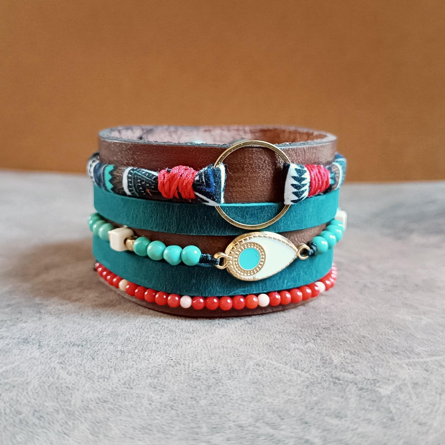 The Warrior Stack Extra Wide Ethnic Leather Cuff. Features dark brown Crazy Horse leather with five layers including red crystal & white howlite beads, natural turquoise beads, a brass evil eye charm, petrol blue leather stripes, and blue ethnic cotton cord.