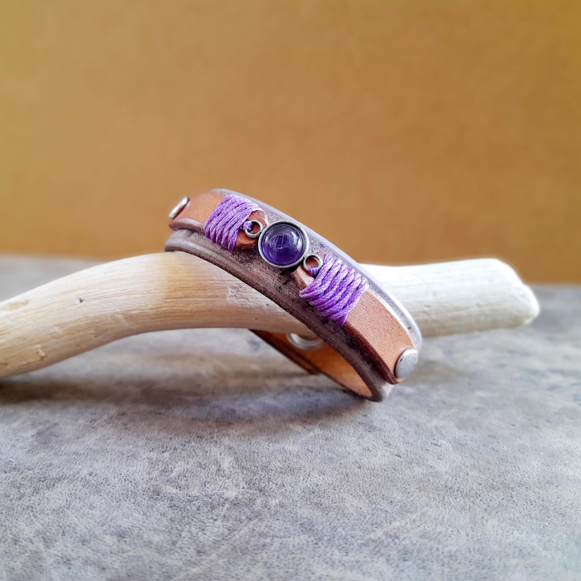 A two-layered leather bracelet with a natural amethyst pendant, featuring purple thread and silver rivets, displayed on a wooden surface.