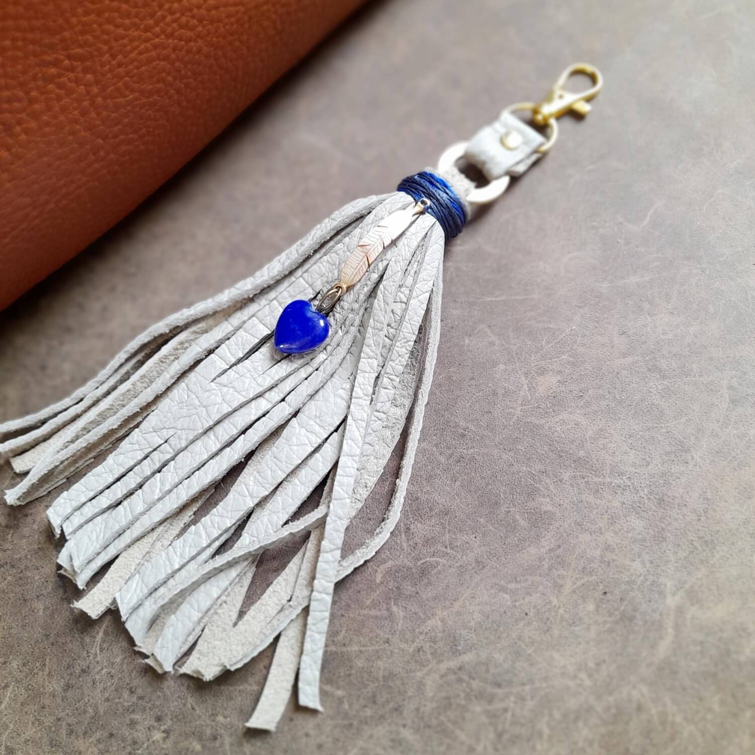 OOAK Lapis Lazuli Heart Charm with a thick gray leather tassel, laid flat. Features a natural Lapis Lazuli heart pendant wrapped in navy blue thread, and a gold-tone brass feather charm.