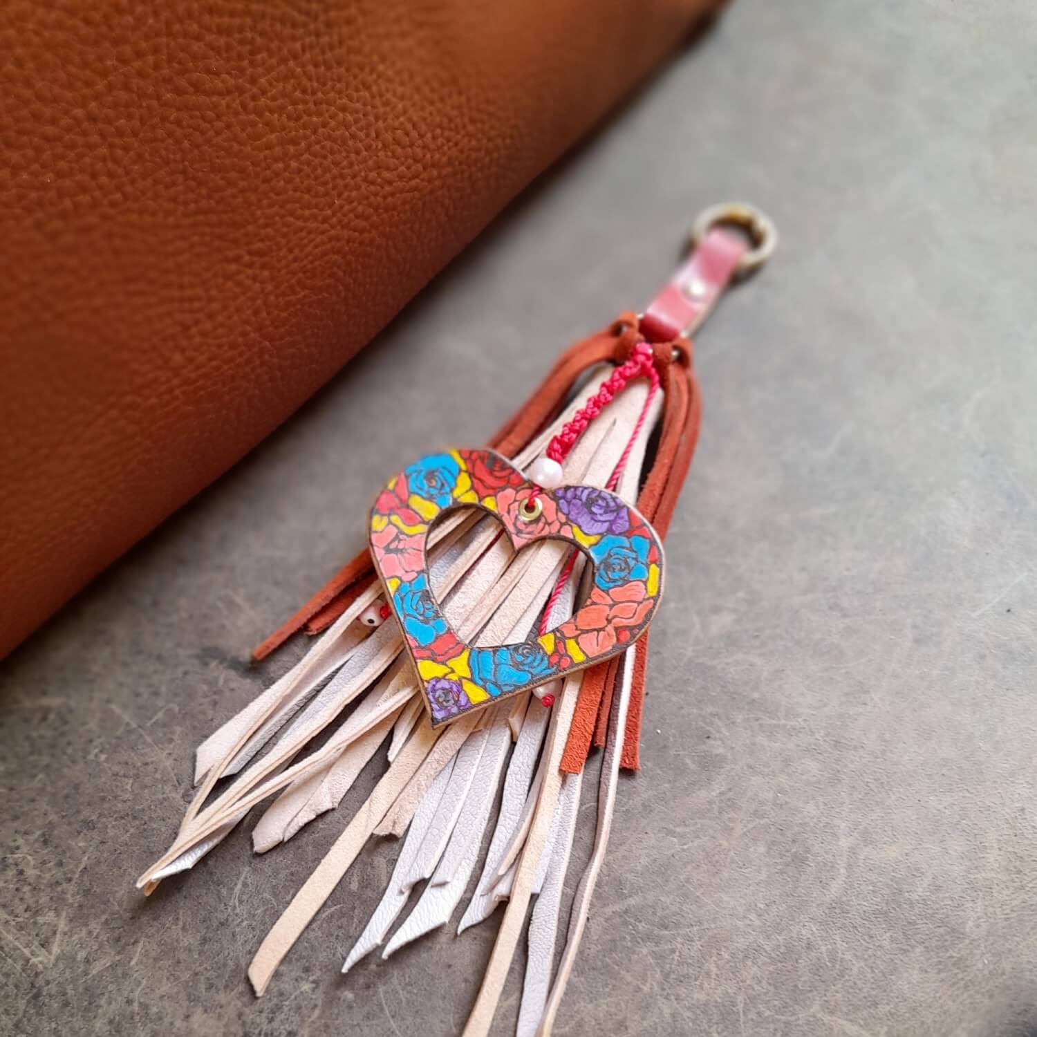 The OOAK Stained Glass Heart Charm: A hand-painted floral leather "hollow heart" with vibrant colors, attached to a flowing pink suede tassel with red fringe. Features a gold-tone O-ring clip.