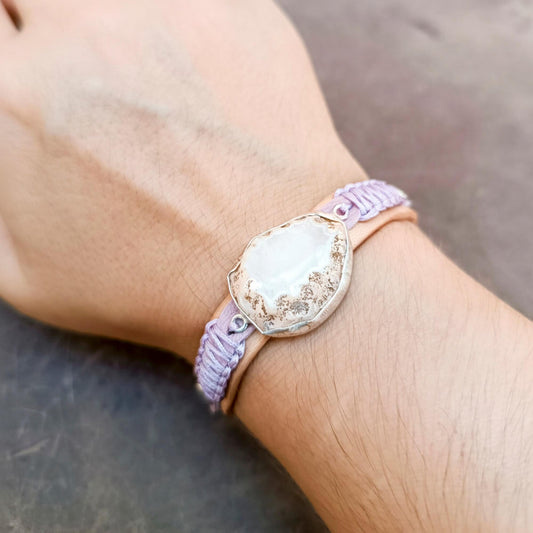 A close-up of a person's wrist wearing a handmade bohemian-style bracelet. The bracelet features a central raw druzy rose quartz stone with a scalloped silver border, wrapped in a light pink leather band and a lavender-purple macrame cord, resting on a dark gray surface.