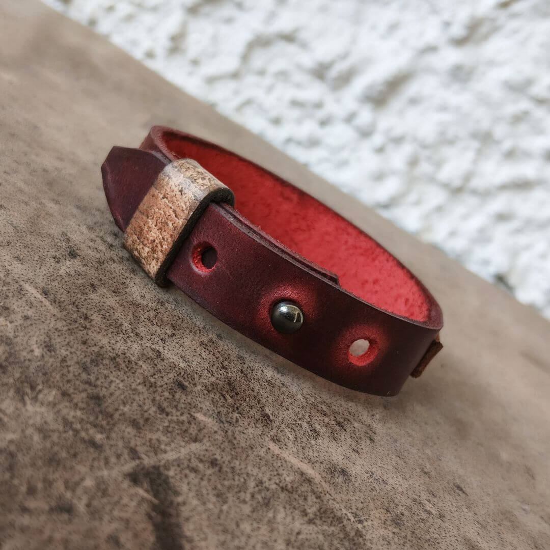 A dark red leather bracelet with a textured tan leather tab and a silver-tone metal rivet, resting on a textured brown surface.