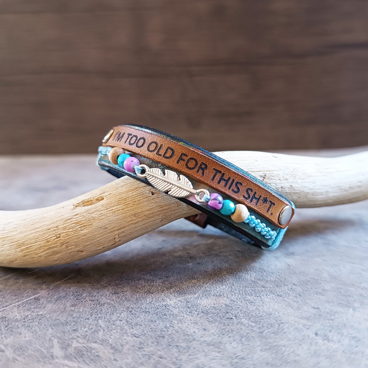 Other side view of The "No BS" Leather Wrap Bracelet. Features a black band hand-dyed with abstract colorful stripes, a thin leather strip laser-engraved with "I'm Too Old for This Sh*t," a row of colorful beads, and a silver-tone feather charm.