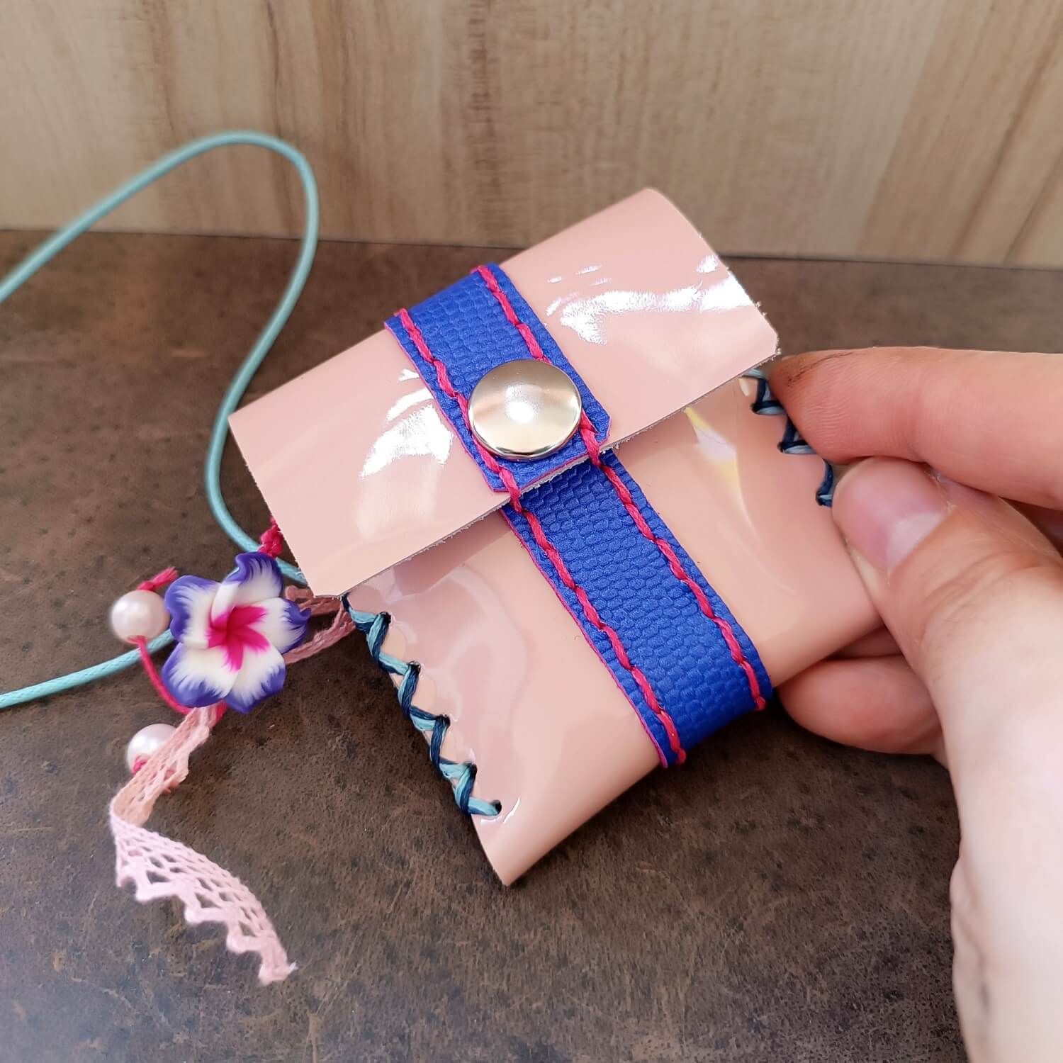 Hand holding a pink pouch with a blue strap and decorative tassel on a wooden surface
