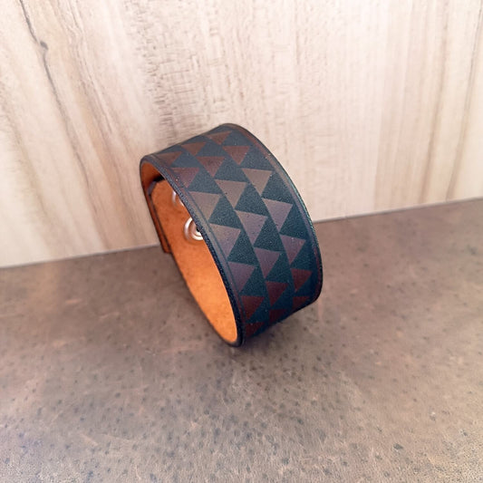 A dark brown leather cuff bracelet with a bold geometric Samoan tribal pattern, displayed against a wooden background.