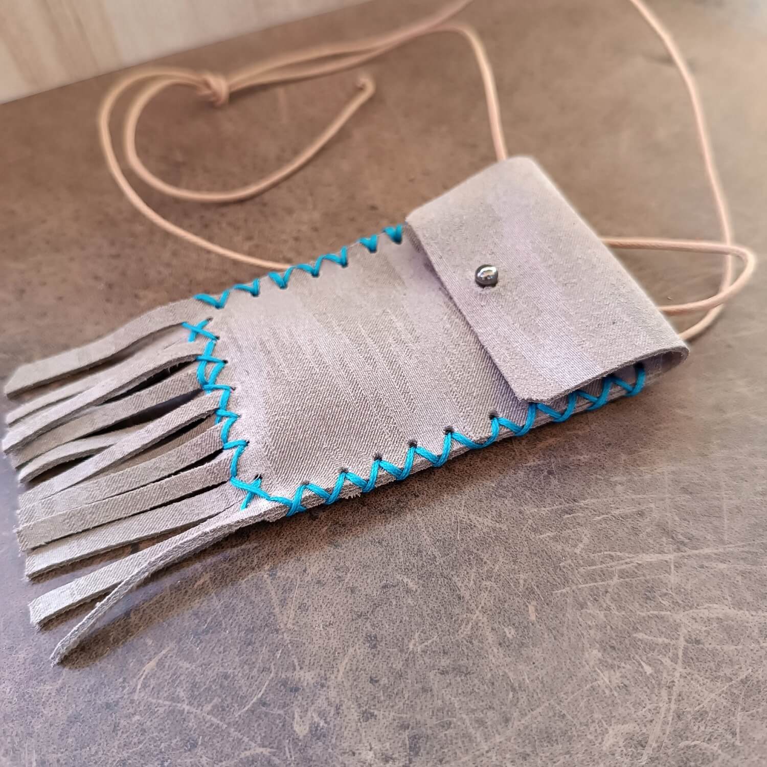Shimmering Gray Leather Neck Bag, crafted from upcycled upholstery leather, with playful fringe and hand-stitched turquoise waxed cord. An organic cotton cord is tied at the top, showing its adjustable length.