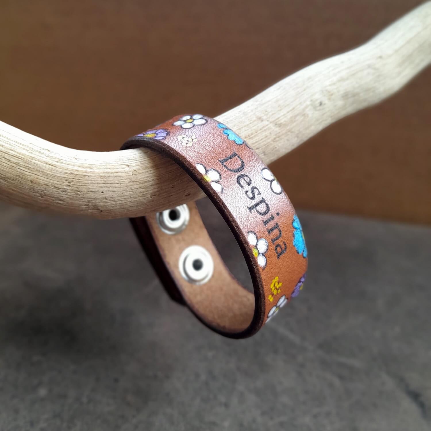 Brown leather bracelet with floral patterns and 'Despina' engraving on a wooden stick.