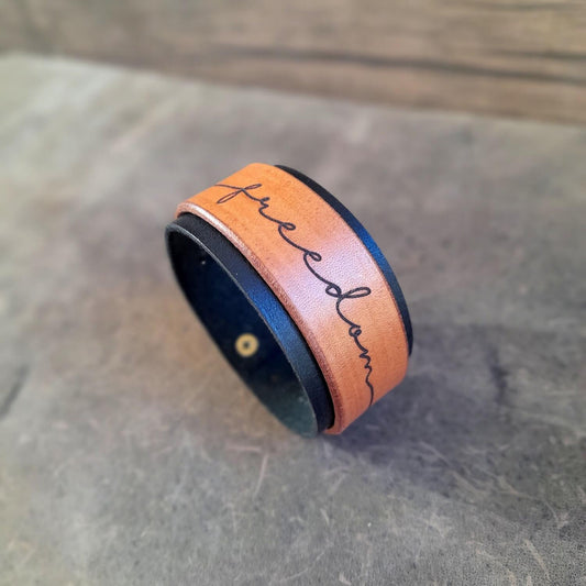 Side view of The Freedom Leather Cuff Bracelet with "Freedom" deeply engraved in calligraphy. Shows the black and brown full grain leather.