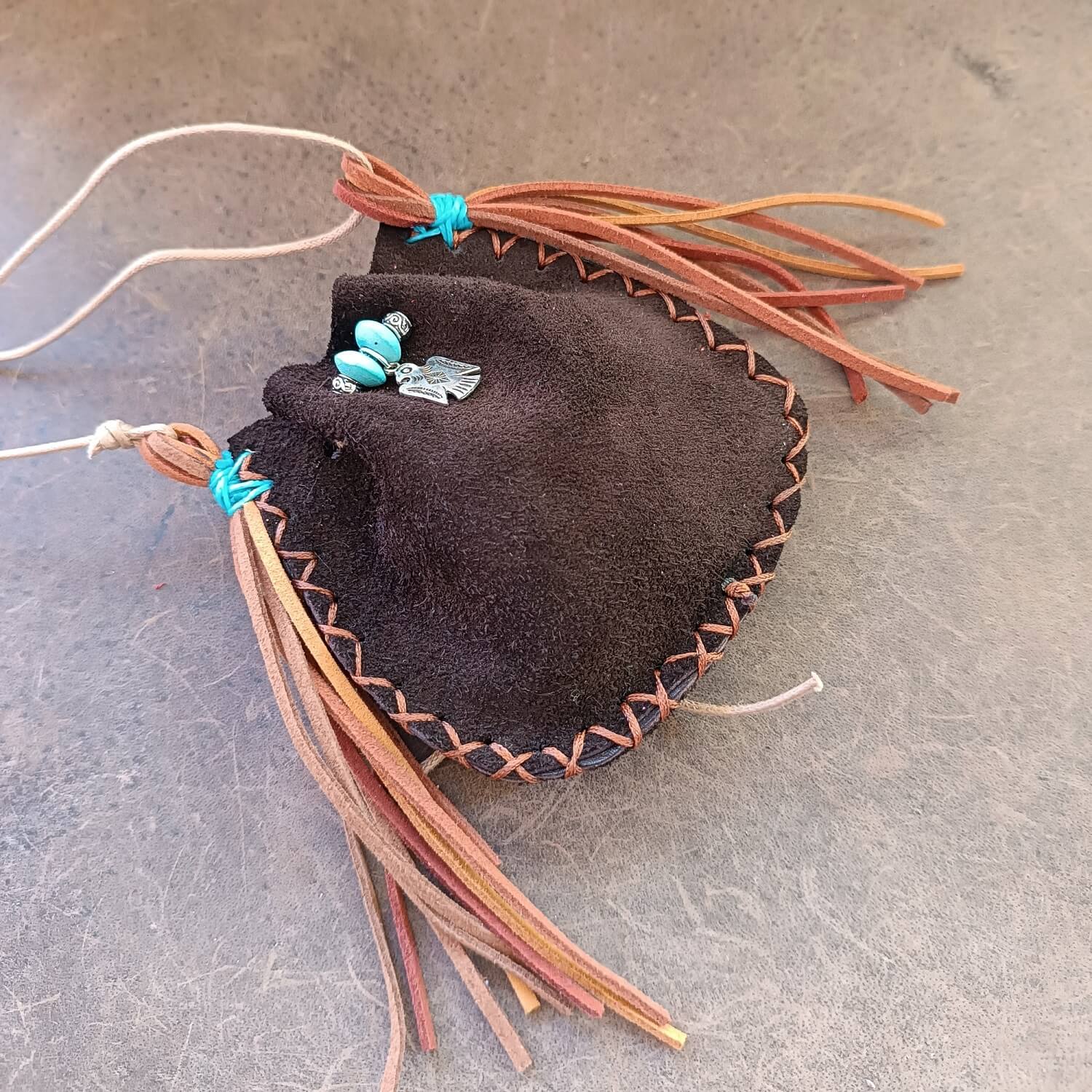 Close-up side view of The Thunderbird Keeper Neck Pouch, highlighting the soft chestnut suede tassels, the silver Thunderbird charm, and the turquoise wooden beads.
