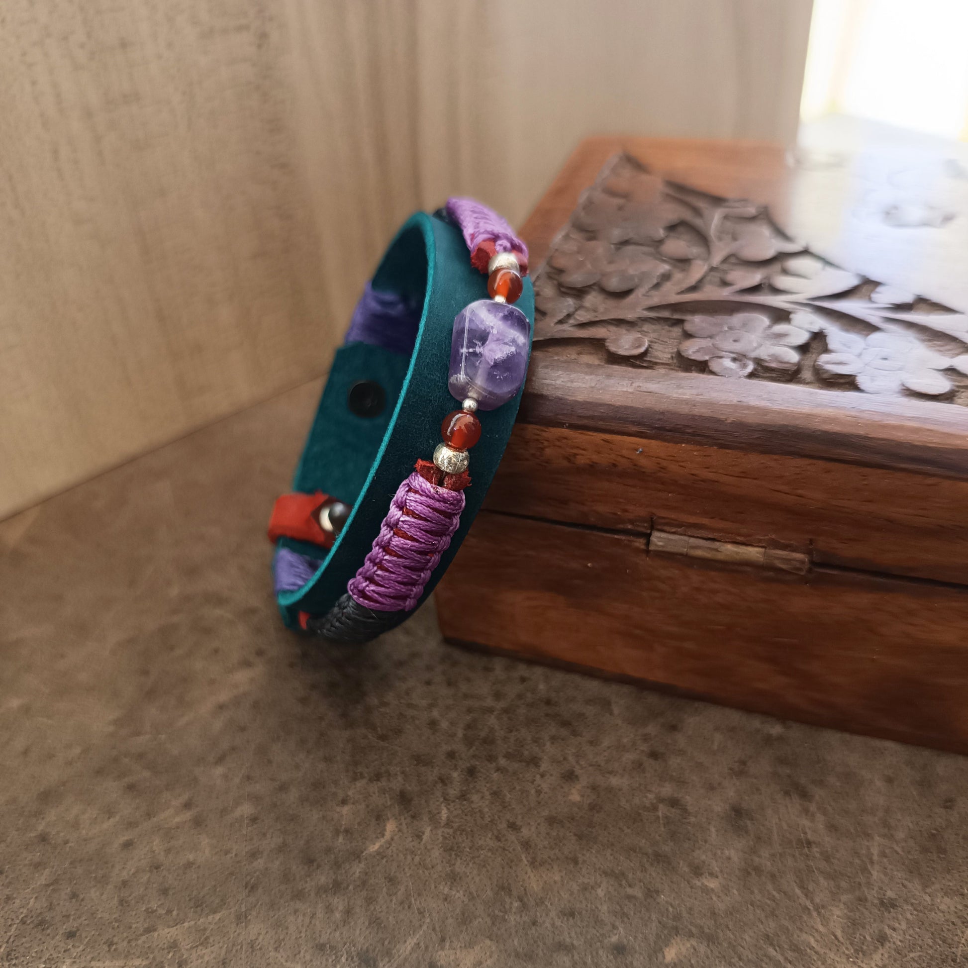 Side view of the amethyst bracelet resting on oak jewelry box, showing braided macramé cord and petrol blue leather