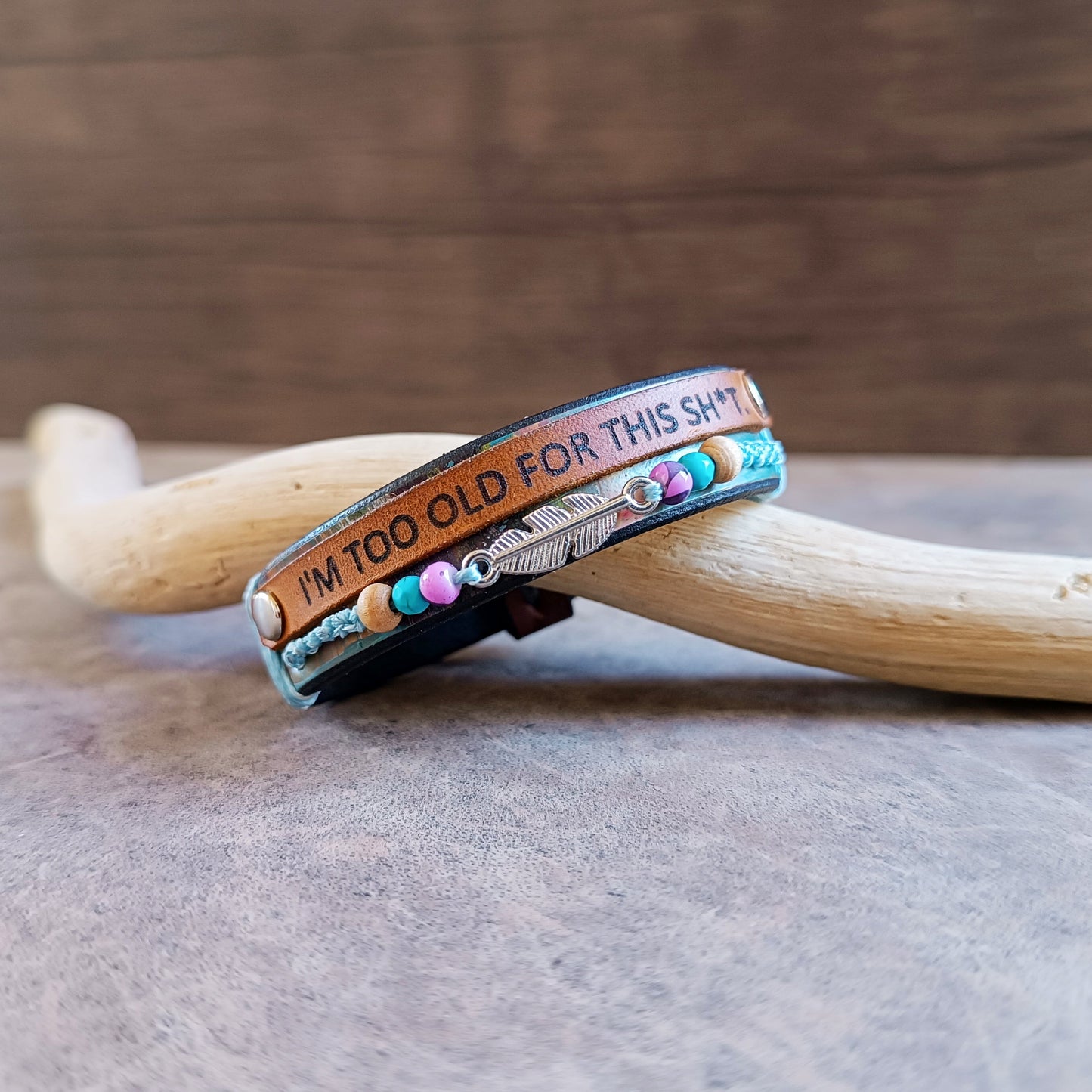 Slight side view of the "No BS" Leather Wrap Bracelet. Features a black band hand-dyed with abstract colorful stripes, a thin leather strip laser-engraved with "I'm Too Old for This Sh*t," a row of colorful beads, and a silver-tone feather charm.