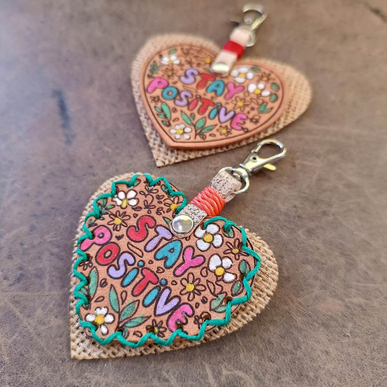 "Stay Positive" Leather Heart Keychains: Two unique, hand-painted hearts made from upcycled leather scraps. One features intricate green hand-stitching, the other is sleek. Both have a jute backing and silver lobster clasp.