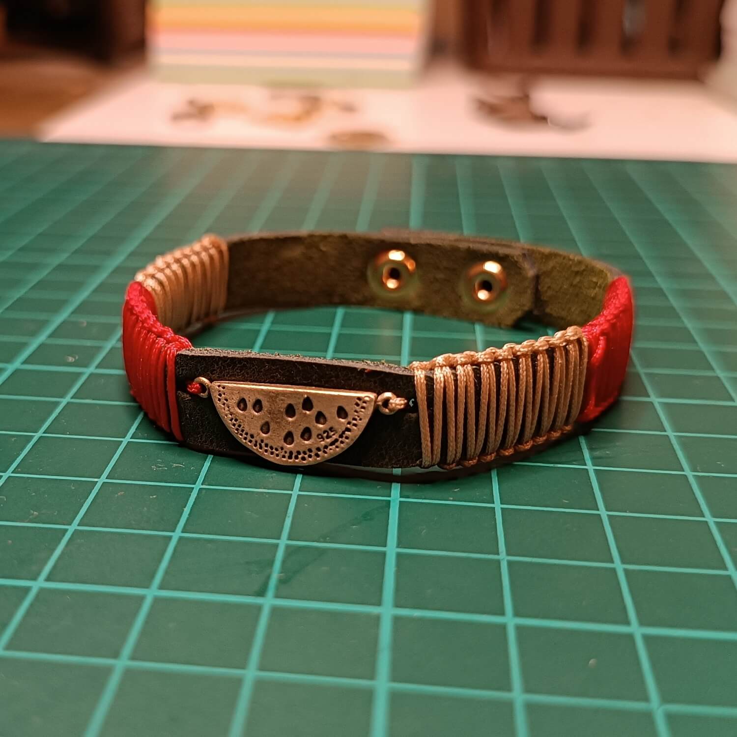 The Sweet Abundance Watermelon Bracelet from the Soulful Echoes Collection, laid flat on my workbench. It features a charming oxidized brass watermelon slice pendant on a deep forest green Crazy Horse leather band, with vibrant red and light bronze macrame cords.