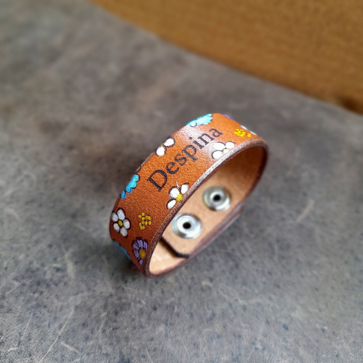 The Bloom & Name Bracelet, a hand-painted floral leather cuff. Shows a custom engraved name on the front, surrounded by small hand-painted flowers, and stainless steel snap buttons.
